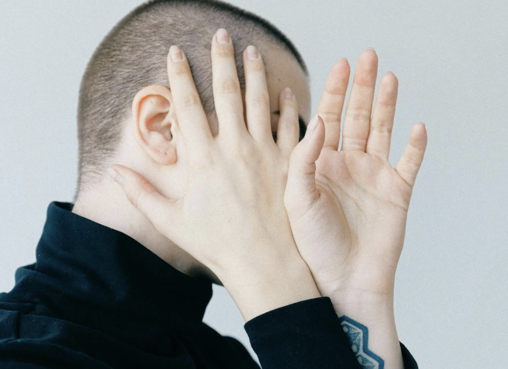 Person with a buzz cut covering their face with their hands, showing a gesture of vulnerability or hesitation, against a plain background, with a tattoo visible on the wrist.