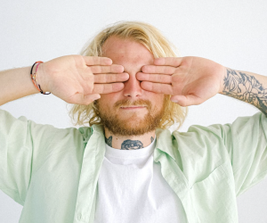 Man with blonde hair and tattoos covering his eyes with his hands, set against a simple light background, conveying a sense of introspection and priva