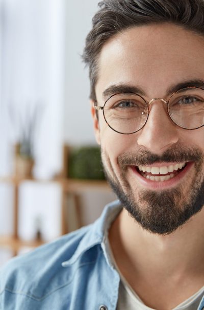 Close up shot of cheerful satisfied attractive male with stubble, has broad smile, wears round spectacles, rejoices success at work, stands against cozy interior. Fashionable designer glad be praised